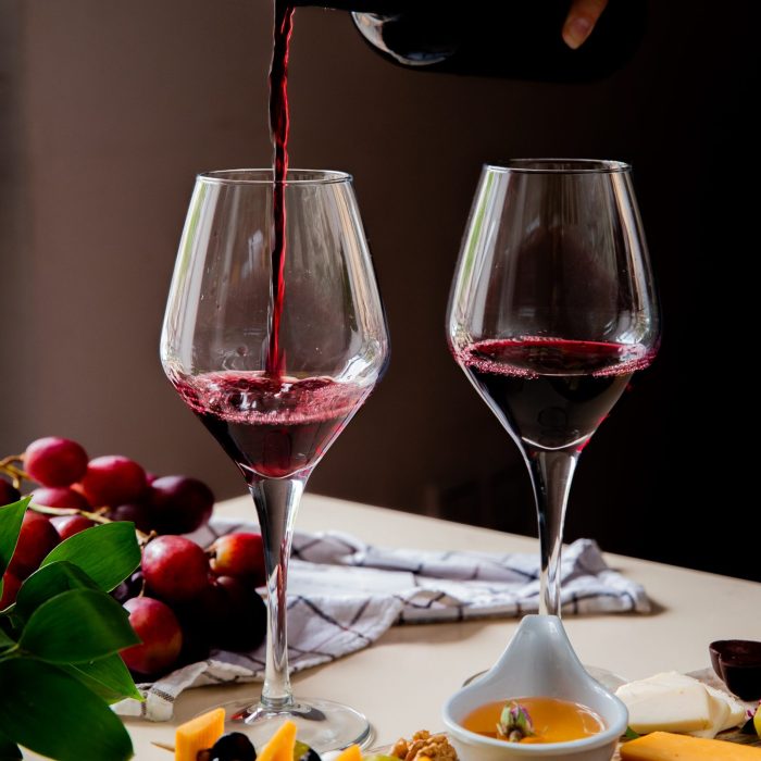 side view of woman hand pouring red wine into glass and different kinds of cheese olive walnut grape on white surface and black background
